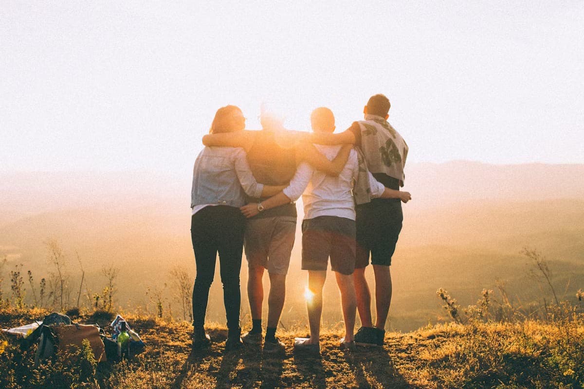 Vier Freund:innen Arm in Arm im Sonnenuntergang, mit Blick über sanfte Hügel — Verbundenheit und Zugehörigkeit.
